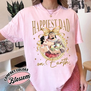 K&ouml;nnte beinhalten: Hellrosa T-Shirt mit goldenem Text "Happiest Dad on Earth". Das Design zeigt eine Cartoon-Illustration eines Vaters und Kindes in einem dekorativen Rahmen. Auf dem Shirt stehen auch die W&ouml;rter "Comfort Colors" und "Blossom".