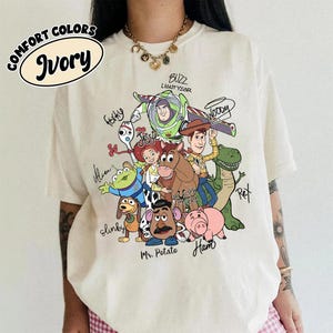 May include: An ivory short-sleeve t-shirt featuring a colorful graphic print of Toy Story characters including Woody, Buzz Lightyear, Jessie, Bullseye, Rex, Slinky Dog, Mr. Potato Head, Ham, Forky, and an Alien, with their names written in black script.