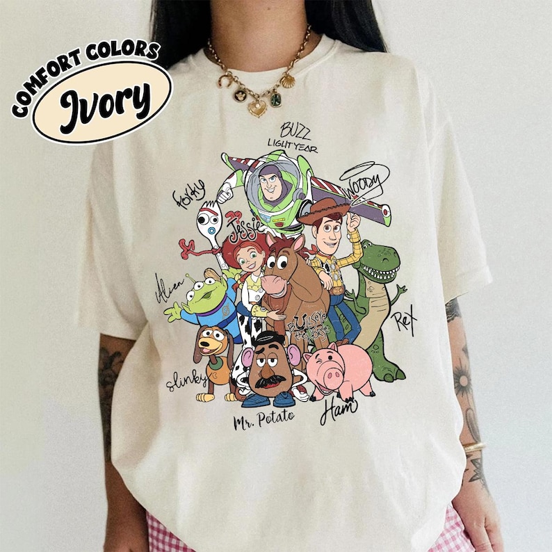 May include: An ivory short-sleeve t-shirt featuring a colorful graphic print of Toy Story characters including Woody, Buzz Lightyear, Jessie, Bullseye, Rex, Slinky Dog, Mr. Potato Head, Ham, Forky, and an Alien, with their names written in black script.