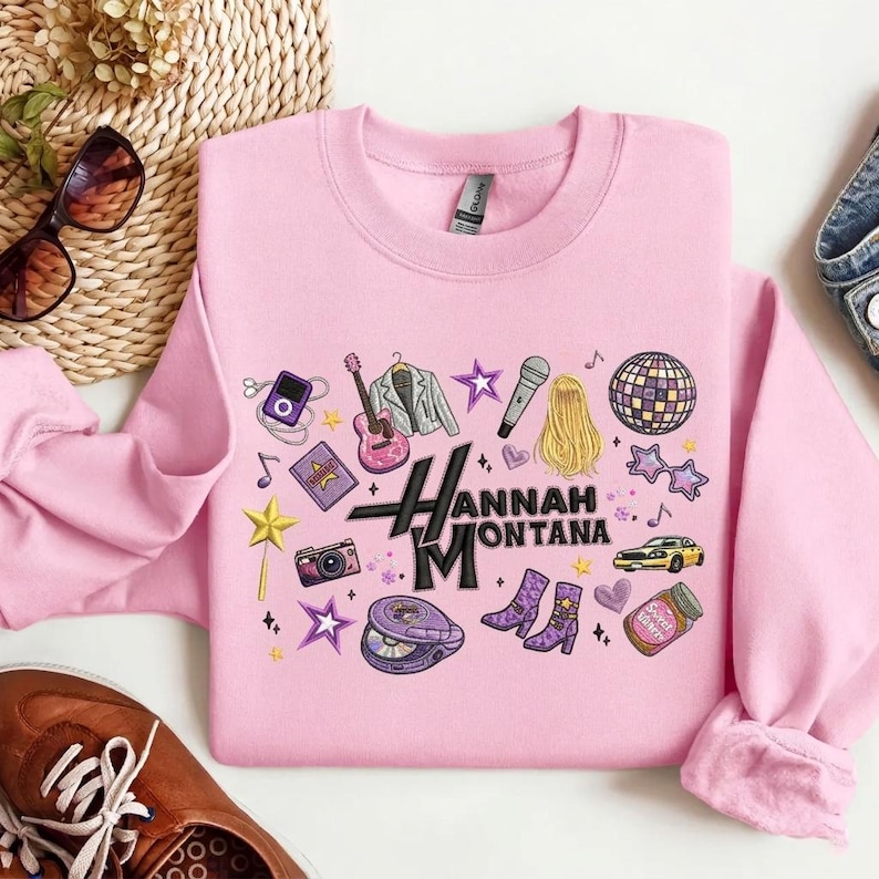May include: Pink sweatshirt with a graphic design featuring a guitar, microphone, stars, and other icons. The text "Hannah Montana" is prominently displayed. The sweatshirt has a crew neck and long sleeves.