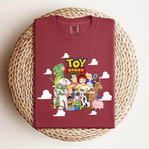 Pu&ograve; includere: Una t-shirt girocollo color marrone scuro con una grafica dei personaggi di Toy Story, tra cui Woody, Buzz Lightyear e Jessie. Il testo recita Toy Story e I Know It I Knew You, circondato da nuvole bianche. La maglietta ha un'etichetta Comfort Colors.