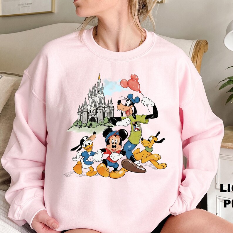 May include: A light pink sweatshirt featuring a colorful graphic of Disney characters Mickey Mouse, Donald Duck, Goofy, and Pluto in front of a castle. The graphic includes red, blue, yellow, and green colors. The sweatshirt is a crew neck style.