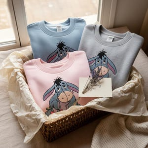 May include: Three crewneck sweatshirts in pastel shades of blue, pink, and grey, each with a cartoon donkey design. They are presented in a woven basket with lavender sprigs and a card that says "thinking of you".