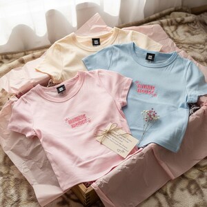 May include: Three pastel-colored t-shirts, pink, light blue, and yellow, are arranged in a wooden box with pink tissue paper. Each shirt has the words "MINNIE MOUSE" embroidered in pink. A small card tied with twine rests on the pink shirt.
