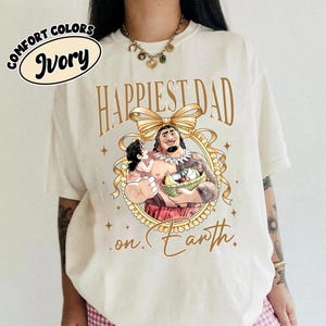K&ouml;nnte beinhalten: Elfenbeinfarbenes T-Shirt mit goldenem Schriftzug "Happiest Dad on Earth". Das Design zeigt eine Cartoon-Illustration eines Mannes, der ein Kind h&auml;lt, umrahmt von einem goldenen Band und Sternen. Die Worte "Comfort Colors" und "Ivory" sind in einem Kreis.