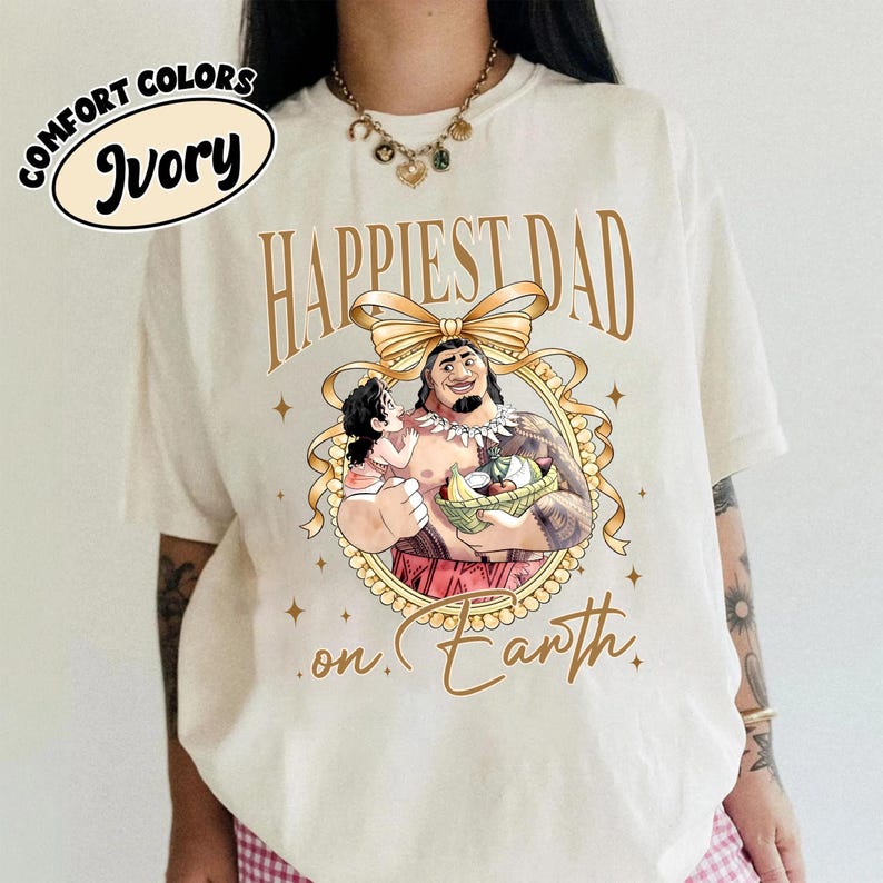 K&ouml;nnte beinhalten: Elfenbeinfarbenes T-Shirt mit goldenem Schriftzug "Happiest Dad on Earth". Das Design zeigt eine Cartoon-Illustration eines Mannes, der ein Kind h&auml;lt, umrahmt von einem goldenen Band und Sternen. Die Worte "Comfort Colors" und "Ivory" sind in einem Kreis.