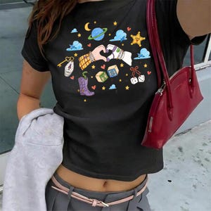 May include: A black short-sleeved t-shirt featuring a colorful graphic print of two hands forming a heart shape, surrounded by illustrations of a planet, stars, clouds, a cowboy boot, dice, alphabet blocks, and a key with the text 1989.