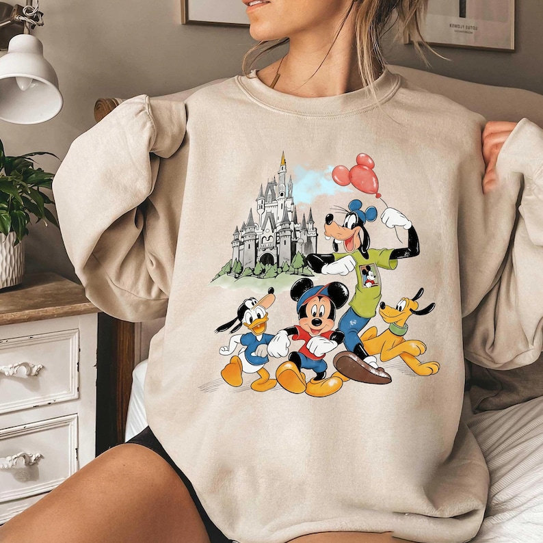 May include: Beige sweatshirt featuring a colorful graphic of Disney characters Mickey Mouse, Donald Duck, Goofy, and Pluto. The design includes a castle and red balloons. The sweatshirt is a neutral color.