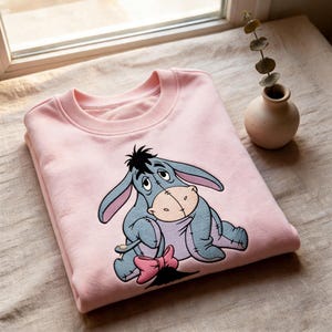 May include: A pale pink sweatshirt with an embroidered Eeyore design. The cartoon donkey is depicted in blue, grey, and pink, with a pink bow. The sweatshirt is folded on a neutral surface, near a window.