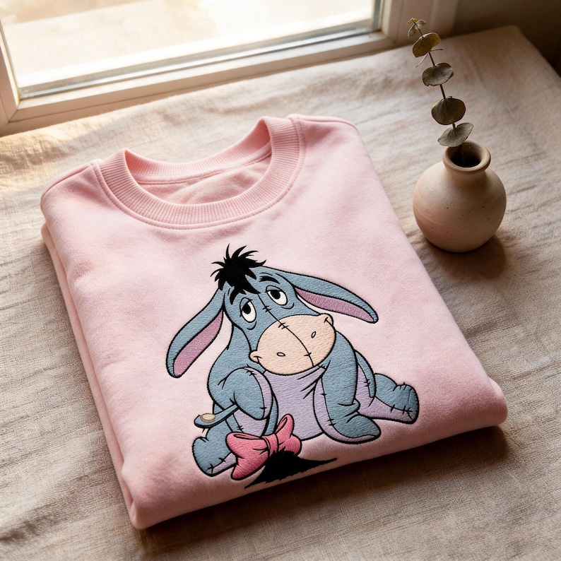 May include: A pale pink sweatshirt with an embroidered Eeyore design. The cartoon donkey is depicted in blue, grey, and pink, with a pink bow. The sweatshirt is folded on a neutral surface, near a window.