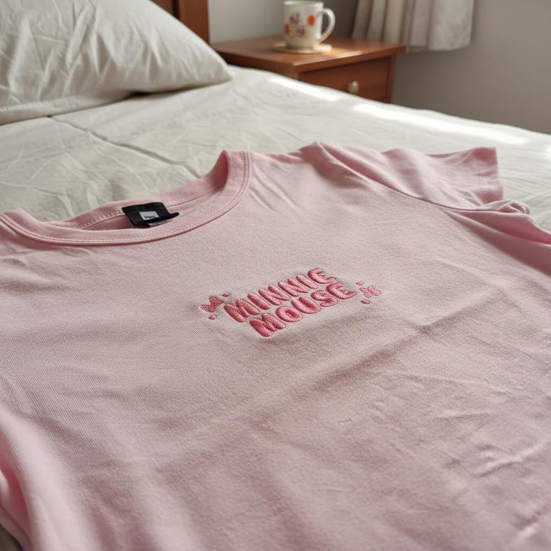 May include: A light pink t-shirt with the embroidered text "MINNIE MOUSE" in a darker pink. The shirt is made of a soft material and has a classic crew neck design. The shirt is laying on a bed.