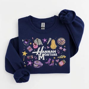 May include: Navy blue sweatshirt with a graphic design featuring the text "Hannah Montana" and various illustrations, including a guitar, microphone, and stars. The sweatshirt is folded.