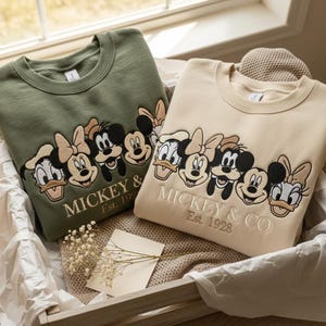 Puede incluir: Dos sudaderas de cuello redondo, una verde oliva y otra beige, con bordados de personajes de Disney. El dise&ntilde;o incluye Mickey Mouse, Minnie Mouse, Donald Duck y Daisy Duck, con el texto "Mickey & Co. Est. 1928".