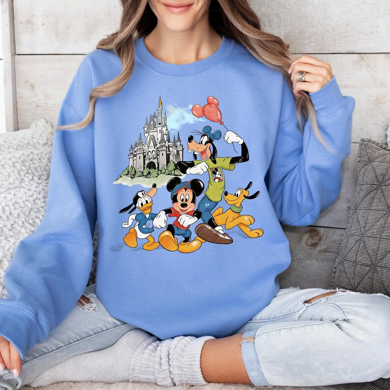 May include: A light blue sweatshirt featuring a graphic of Mickey Mouse, Donald Duck, Goofy, and Pluto in front of a castle. Mickey Mouse is wearing red shorts and a black hat. Goofy holds red balloons. The sweatshirt is long-sleeved.