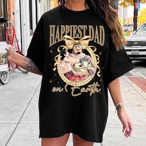 K&ouml;nnte beinhalten: Schwarzes T-Shirt mit einem goldenen Aufdruck "Happiest Dad on Earth". Die Grafik zeigt eine Cartoon-Illustration eines Mannes, der ein Kind und einen Obstkorb h&auml;lt, umgeben von einem goldenen Rahmen und einer Schleife.