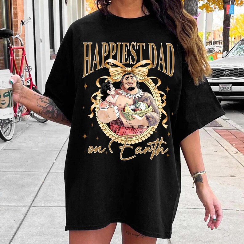 K&ouml;nnte beinhalten: Schwarzes T-Shirt mit einem goldenen Aufdruck "Happiest Dad on Earth". Die Grafik zeigt eine Cartoon-Illustration eines Mannes, der ein Kind und einen Obstkorb h&auml;lt, umgeben von einem goldenen Rahmen und einer Schleife.