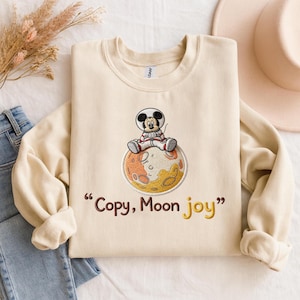May include: Cream-colored sweatshirt featuring an embroidered design of a cartoon mouse in a spacesuit sitting on a planet. The text "Copy, Moon joy" is embroidered below the planet in brown and yellow. The sweatshirt is folded on a surface with jeans and a hat.