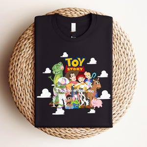 Pu&ograve; includere: Una t-shirt nera a girocollo con una grafica colorata dei personaggi di Toy Story, tra cui Woody, Buzz Lightyear, Jessie e Rex. Il testo "TOY STORY" appare in giallo e rosso, con la frase "I Knew It I Knew You" scritta al di sotto.