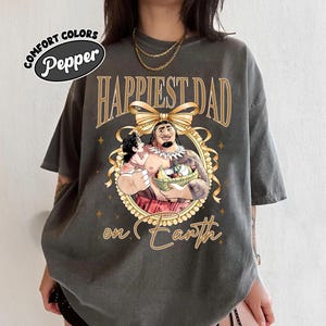 K&ouml;nnte beinhalten: Dunkelgraues T-Shirt mit dem goldenen Schriftzug "Happiest Dad on Earth". Das Shirt zeigt eine Cartoon-Illustration eines Mannes, der ein Kind und einen Korb mit Essen h&auml;lt, umrahmt von einer goldenen Schleife und einem dekorativen Rand. Das Shirt tr&auml;gt auch den Text "Comfort Colors Pepper".