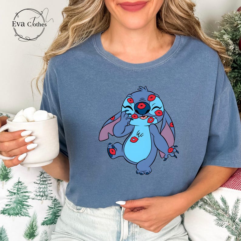 Puede incluir: Camiseta azul con un personaje de dibujos animados cubierto de marcas de besos rojos. El personaje es azul con orejas grandes y una barriga azul claro. La camiseta tiene el texto "Eva Clothes" en un logotipo circular.