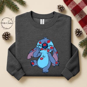 Puede incluir: Sudadera gris oscuro con un personaje de dibujos animados azul cubierto de marcas de besos rojos. El personaje tiene orejas grandes y est&aacute; sonriendo. La sudadera tiene cuello redondo y mangas largas. La etiqueta de la marca es visible en el interior del cuello.