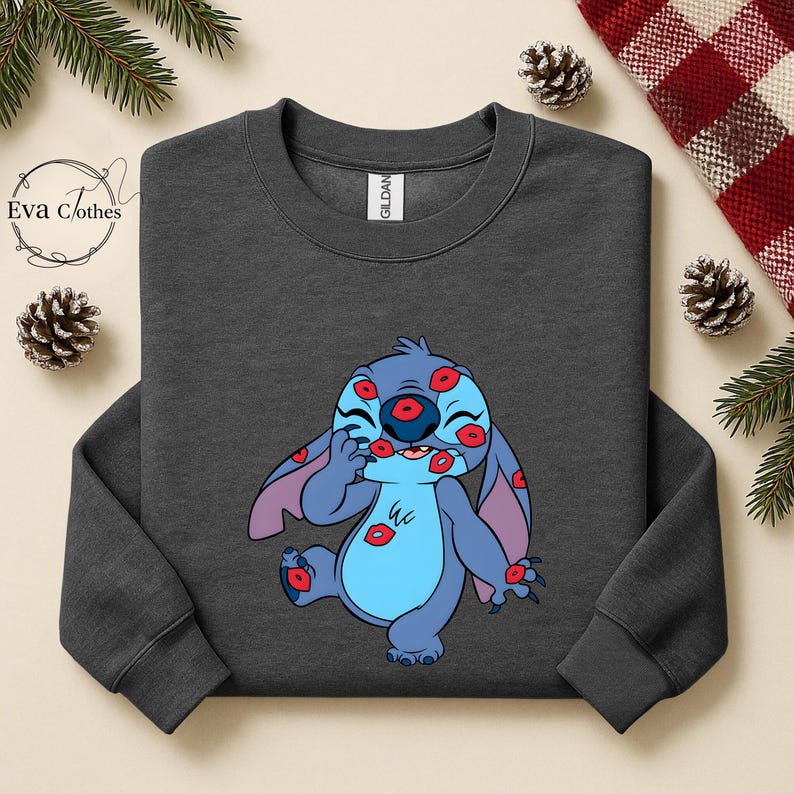 Puede incluir: Sudadera gris oscuro con un personaje de dibujos animados azul cubierto de marcas de besos rojos. El personaje tiene orejas grandes y est&aacute; sonriendo. La sudadera tiene cuello redondo y mangas largas. La etiqueta de la marca es visible en el interior del cuello.
