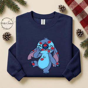 Puede incluir: Sudadera azul marino con un personaje de dibujos animados cubierto de marcas de besos rojos. El personaje es azul con orejas grandes y una expresi&oacute;n juguetona. La sudadera tiene cuello redondo y la marca "Gildan" es visible.