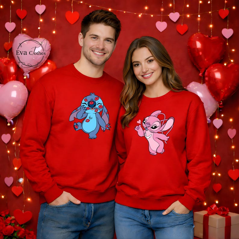 Puede incluir: Dos personas con sudaderas rojas a juego. Una sudadera presenta un personaje de dibujos animados azul cubierto de besos rojos, y la otra tiene un personaje de dibujos animados rosa sosteniendo un coraz&oacute;n. El fondo incluye globos en forma de coraz&oacute;n y luces de cadena.