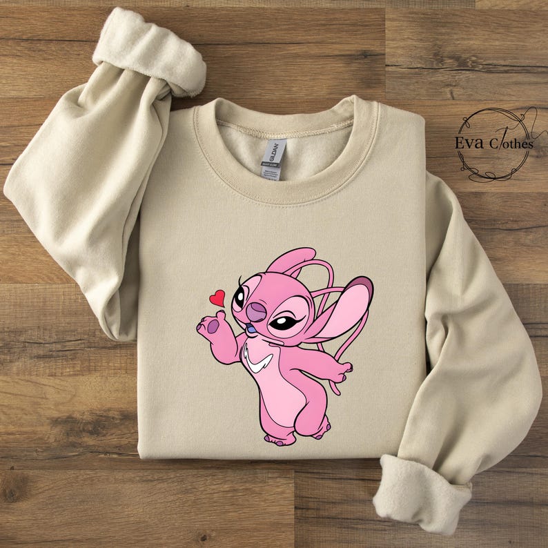 Puede incluir: Sudadera beige con un personaje de dibujos animados rosa sosteniendo un coraz&oacute;n. El personaje es rosa con orejas grandes y un dise&ntilde;o en forma de coraz&oacute;n. La sudadera tiene mangas largas con pu&ntilde;os enrollados. El texto "Eva Clothes" est&aacute; en la esquina superior derecha.