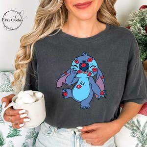 Puede incluir: Camiseta gris oscuro con un personaje de dibujos animados cubierto de besos de l&aacute;piz labial rojo. El personaje es azul con orejas grandes y una barriga azul claro. La camiseta tiene el logo de Eva Clothes.