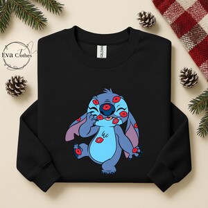 Puede incluir: Sudadera negra con un personaje de dibujos animados azul cubierto de marcas de besos rojos. El personaje tiene orejas grandes y est&aacute; sonriendo. La sudadera tiene cuello redondo y mangas largas. La marca "Gildan" es visible en la etiqueta.