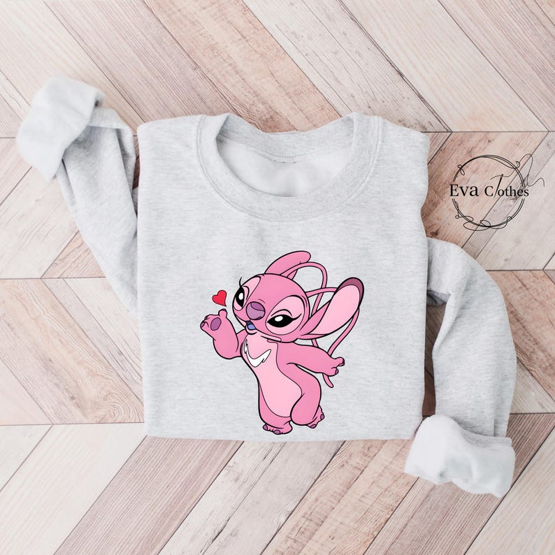 Puede incluir: Sudadera gris claro con un personaje de dibujos animados rosa soplando un beso y sosteniendo un coraz&oacute;n. El personaje tiene orejas grandes. La sudadera tiene mangas largas y el texto "Eva Clothes" es visible.