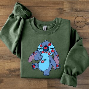 Puede incluir: Una sudadera verde oliva con un personaje de dibujos animados cubierto de huellas de labios rojos. El personaje es azul con orejas grandes y una barriga azul claro. La sudadera tiene mangas largas y la marca "Eva Clothes" es visible en la esquina superior derecha.