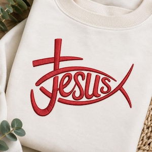 May include: A cream-colored crewneck sweatshirt featuring a red embroidered design on the front. The embroidery depicts a stylized fish shape integrated with a cross and the word Jesus written in a cursive font within the center of the fish.