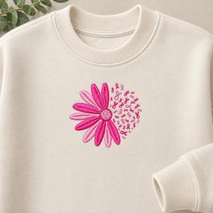 Könnte beinhalten: Ein cremefarbenes Sweatshirt mit Rundhalsausschnitt und einer Stickerei auf der Brust. Das Motiv besteht aus einer Blume mit rosa und hellrosa Blütenblättern auf der linken Seite, während die rechte Seite aus vielen kleinen rosa Schleifen besteht.