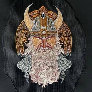 May include: An embroidered patch on a black fabric base featuring a detailed Viking warrior with a long beard and a horned helmet. The design uses gold, silver, and white thread to depict intricate knotwork patterns and a central runic symbol.