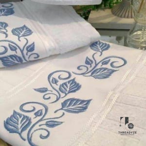 Puede incluir: Toallas de baño blancas con diseños bordados en azul de hojas y vides. Las toallas tienen un borde decorativo y están dobladas. El bordado es detallado y añade un toque de elegancia a la decoración del baño.