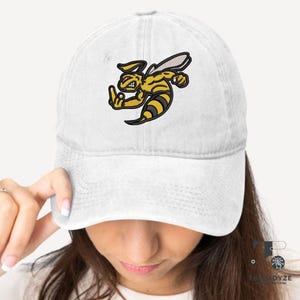 May include: White baseball cap featuring an embroidered cartoon bee in yellow and black, giving the middle finger. The bee has a stern expression. The cap is made of a soft, faded fabric.