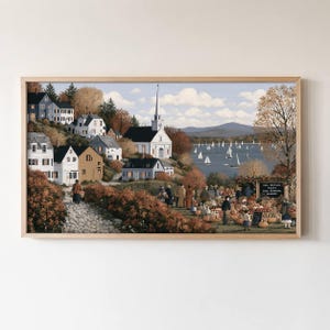 May include: A framed landscape painting depicting a coastal village with white houses, a church, and a harbor filled with sailboats. In the foreground, people gather at a fall festival with pumpkins and a sign reading Fall Festival Today Cider Pumpkins Hayrides.