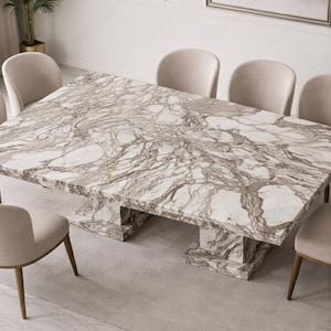 May include: A rectangular dining table with a marble top and a pedestal base. The table is surrounded by six upholstered chairs. The marble top has a white and grey veined pattern. The chairs are a light beige colour. The table is approximately 240 cm long.