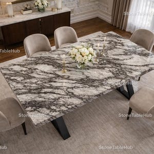 May include: A rectangular marble dining table with a black base, surrounded by six beige upholstered chairs. The table has a white and gray veined pattern. A vase of white flowers and candles adorn the table. The room has a neutral color palette.