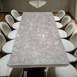 Puede incluir: Una mesa de comedor rectangular con una tapa de piedra rosa y blanca, rodeada de sillas blancas. La superficie de la mesa tiene un patrón de mosaico. Una lámpara de araña de cristal cuelga sobre la mesa. Las sillas tienen asientos blancos y respaldos color crema.