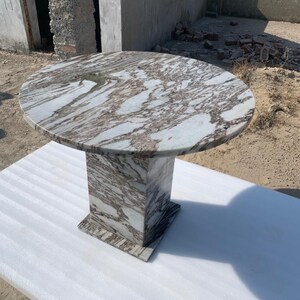 Op de afbeelding: Een ronde marmeren tafel met een voetstuk. Het tafelblad heeft een wervelend patroon van wit en bruin. De basis is een vierkante kolom. De tafel staat op een wit oppervlak.