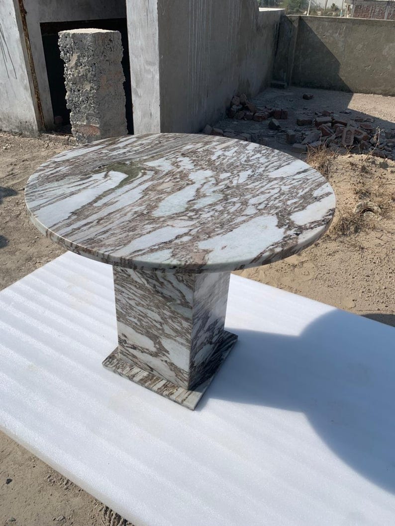 Op de afbeelding: Een ronde marmeren tafel met een voetstuk. Het tafelblad heeft een wervelend patroon van wit en bruin. De basis is een vierkante kolom. De tafel staat op een wit oppervlak.