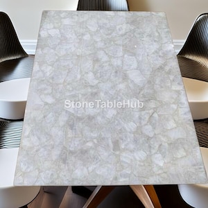 Puede incluir: Una mesa rectangular de piedra gris claro con una superficie texturizada única. La mesa está sostenida por patas de madera y rodeada por cuatro sillas modernas con asientos blancos y respaldos negros y plateados. El texto "StoneTableHub" es visible en la mesa.