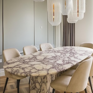 May include: An oval marble dining table with a unique veined pattern, supported by a matching pedestal base. Beige upholstered chairs with black legs surround the table. Crystal chandeliers hang above the table, illuminating the dining area.