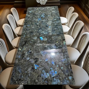 May include: A large rectangular dining table with a dark, patterned stone top. The table is surrounded by white upholstered chairs. A crystal chandelier hangs above. The table's surface has blue and gray hues, with the text "StoneTableHub" visible.