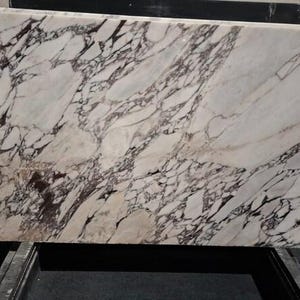 May include: A rectangular slab of marble with a white background and intricate dark brown veining patterns. The stone is displayed horizontally, showcasing its natural variations and textures. This marble slab is suitable for countertops or decorative surfaces.