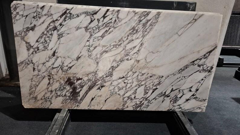 May include: A rectangular slab of marble with a white background and intricate dark brown veining patterns. The stone is displayed horizontally, showcasing its natural variations and textures. This marble slab is suitable for countertops or decorative surfaces.