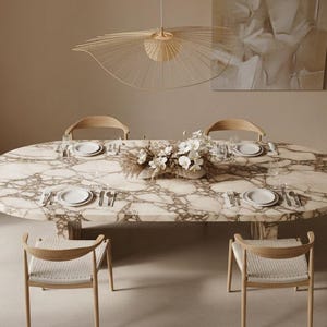 Op de afbeelding: Een ovale marmeren eettafel, gedekt voor zes personen met witte borden, bestek en glazen. Vier houten stoelen met geweven zittingen staan rond de tafel. Een decoratief bloemstuk en een unieke lamp maken de elegante setting compleet.